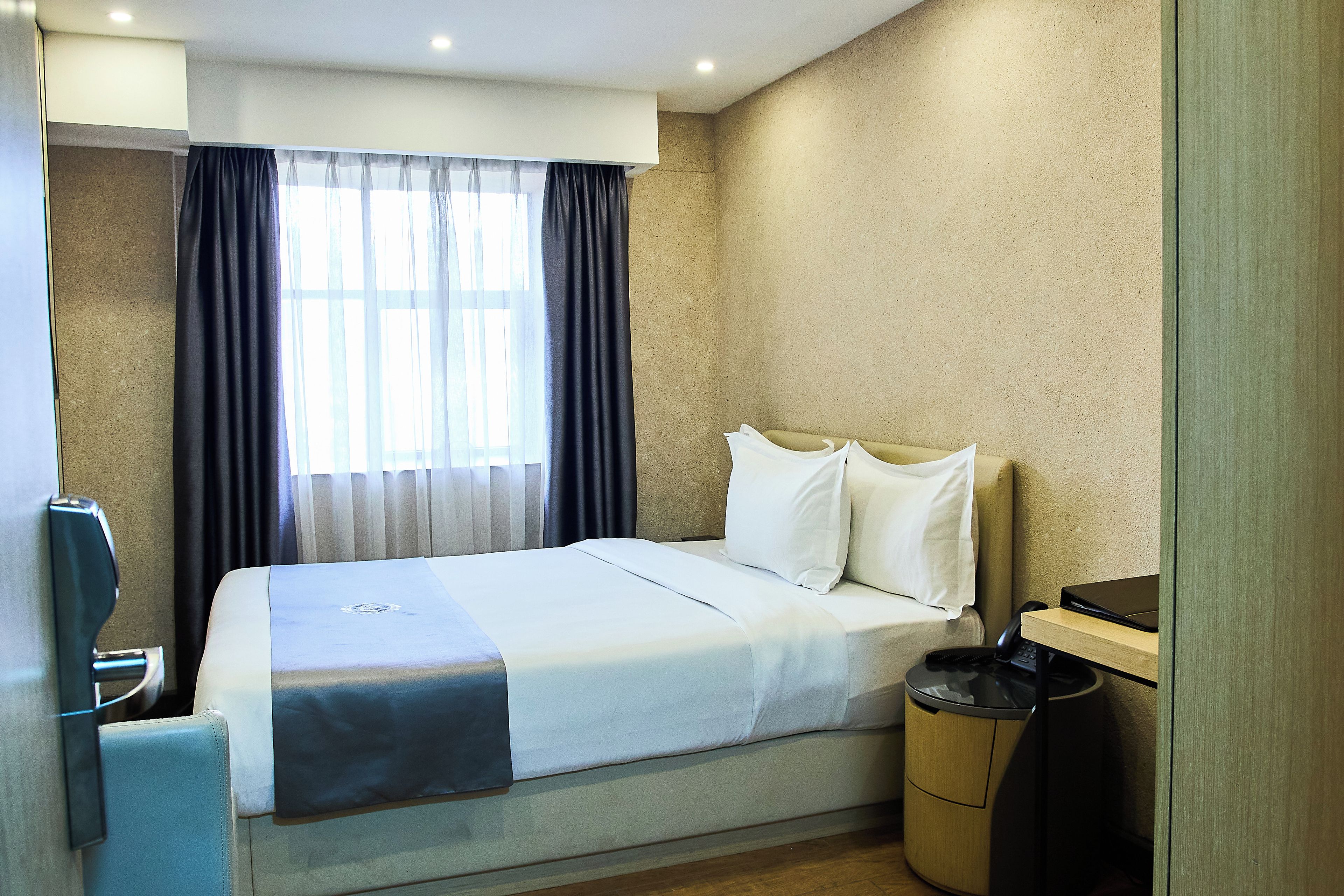 A hotel room with a bed, a bedside table, a lamp, a window with curtains, and a trash bin.