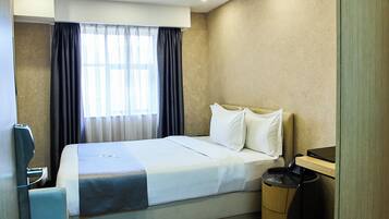 A hotel room with a bed, a bedside table, a lamp, a window with curtains, and a trash bin.