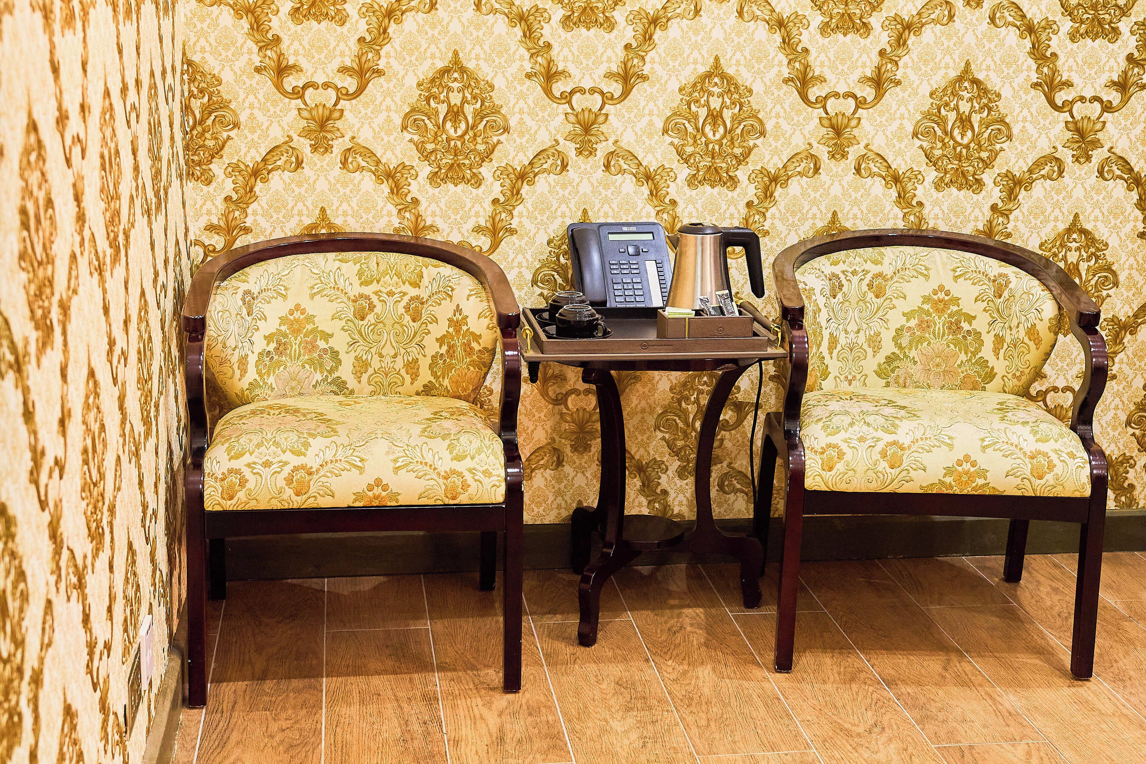 Dos sillones tapizados con estampado floral, una mesita con un teléfono y una cafetera, y un papel tapiz de fondo con motivos.
