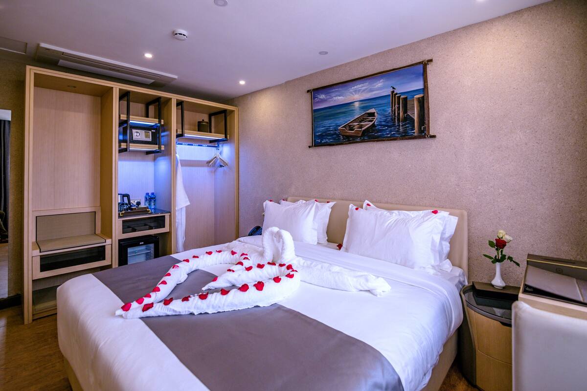 A hotel room with a neatly made bed, a painting of a boat on water, and a minibar.