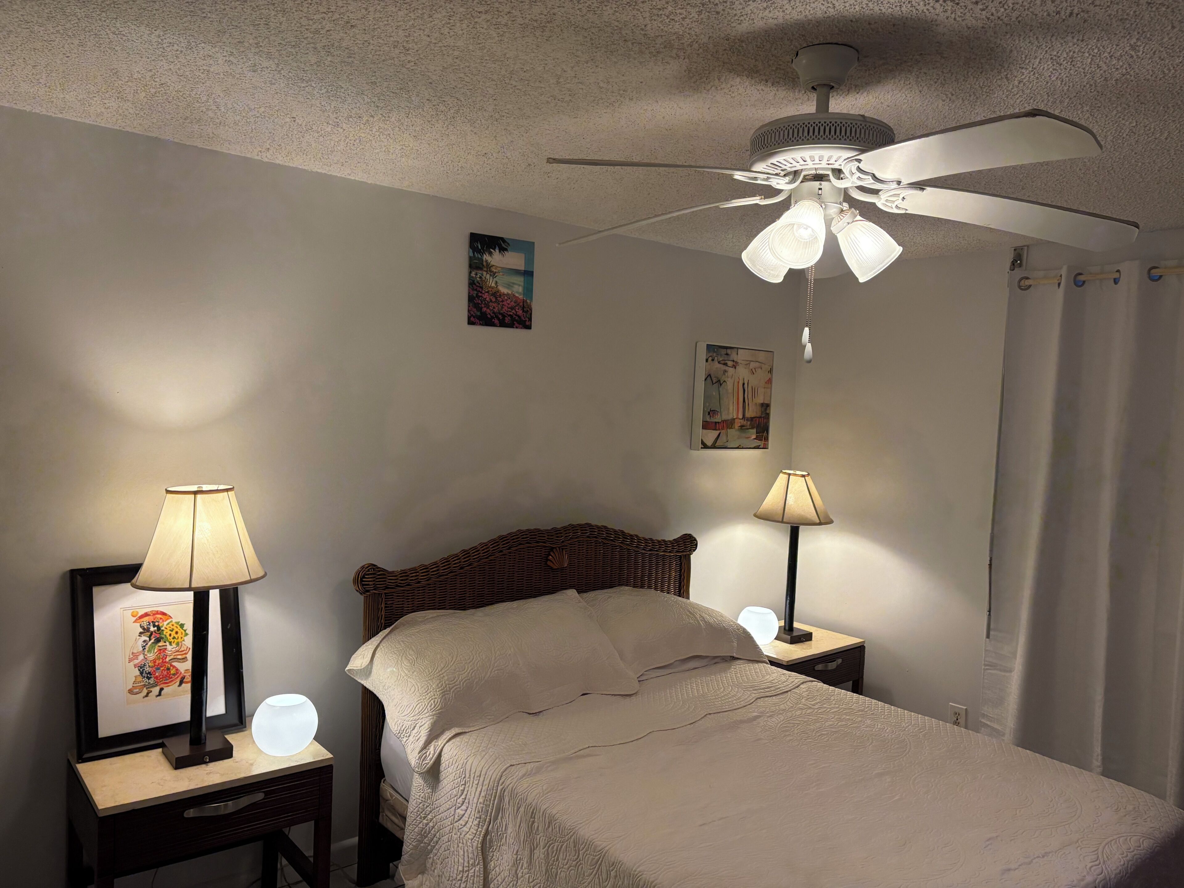 A bedroom with a bed, two bedside tables with lamps, a ceiling fan, and a painting on the wall.