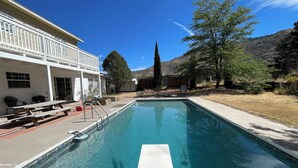 Une piscine à l’eau d’un bleu cristallin, entourée d’un patio aménagé avec du mobilier d’extérieur, un bâtiment blanc doté de grandes fenêtres et un paysage montagneux en arrière-plan.