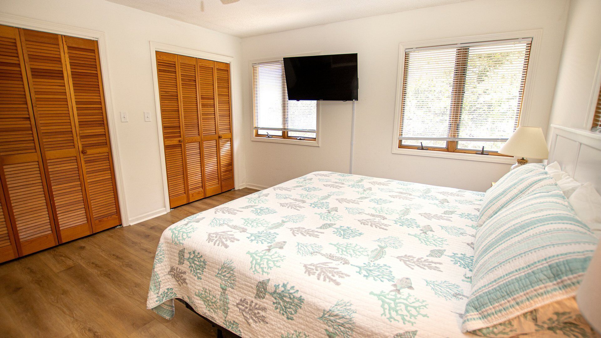 A bedroom with a bed, wooden closet, a TV mounted on the wall, and two windows with blinds.