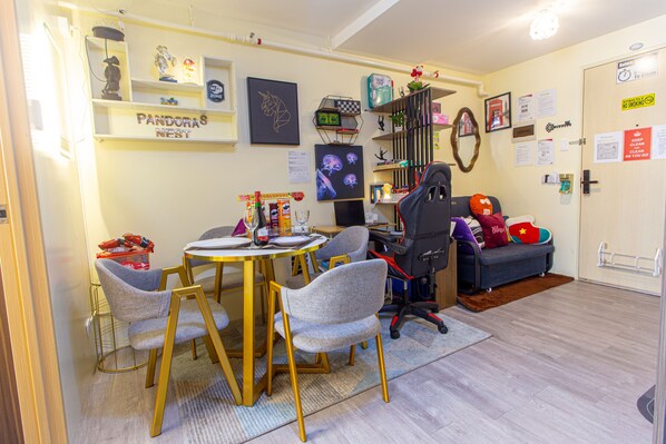 A compact living space with a dining area, a gaming chair, a sofa with colorful cushions, and a bookshelf. - Pandora’s Nest Pasig 2BR Family & Pet-Friendly Haven for Extended Stays (Pasig)