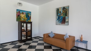 A room with a checkered floor, a brown sofa with patterned pillows, a small table with a bottle, and two paintings on the walls.