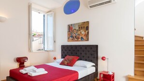 A bedroom with a large bed, a painting on the wall, a window, and a red side table. - Banchi Suite - Castel Sant'Angelo (Rome)