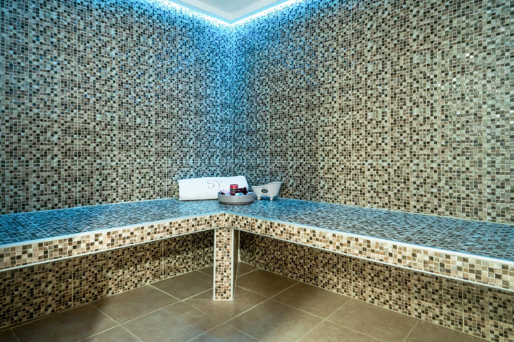 A small, enclosed bathroom with mosaic tile walls and floor, a sink, and a mirror.