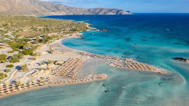 Vue aérienne d’une zone côtière avec une plage, des eaux d’un bleu limpide et un paysage montagneux en arrière-plan.