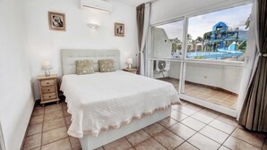 A hotel room with a bed, bedside tables, a large window with a view of a water park, and an air conditioning unit.