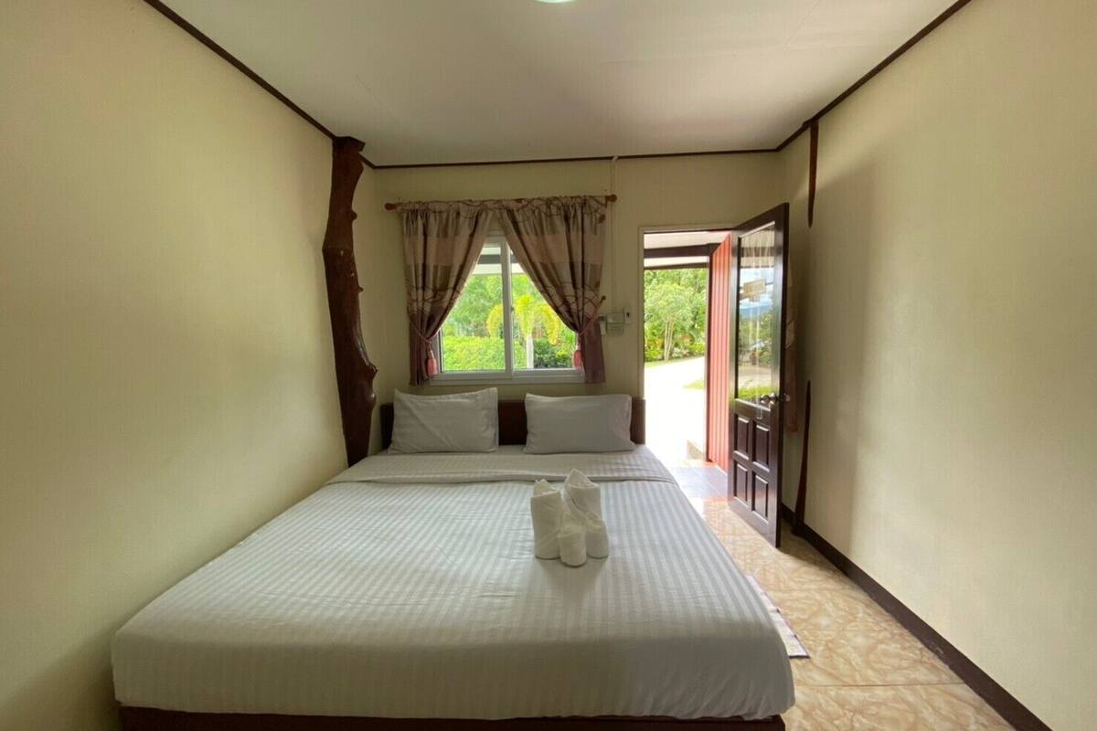 Cama bem arrumada com lençóis e almofadas brancos, cabeceira de madeira e janela com cortinas.
