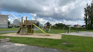 A playground with a slide and swings, surrounded by grass and a few buildings in the background. - Chouette Appartement 5 Personnes (De Haan)