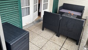 A small outdoor seating area with wicker furniture and a table, a door with a number sign, and a white brick wall. - Chouette Appartement 5 Personnes (De Haan)
