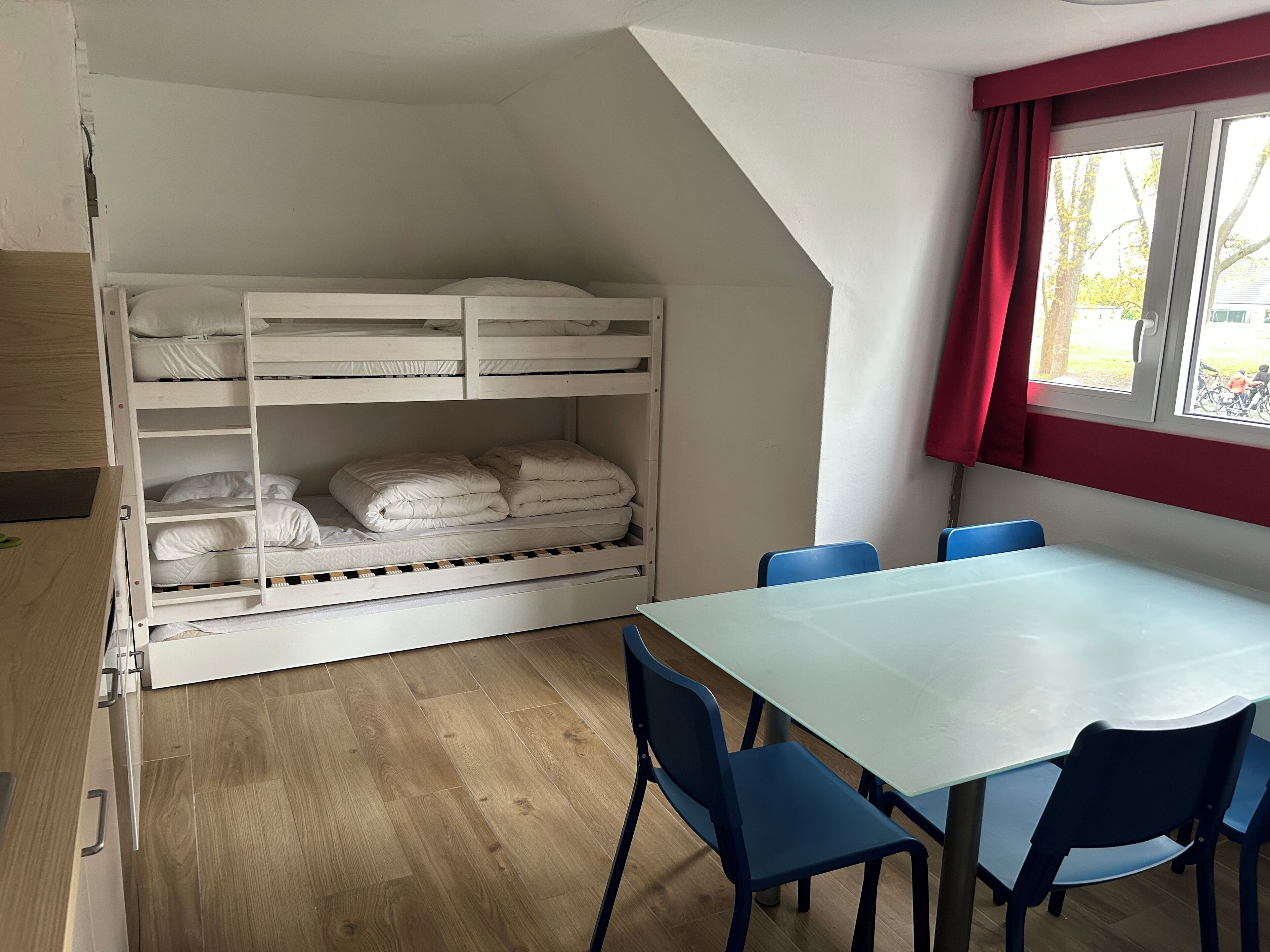 A room with a bunk bed, a table with chairs, and a window with red curtains.