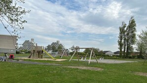 A playground with swings and a slide, surrounded by grass and trees. - Chouette Appartement 5 Personnes (De Haan)