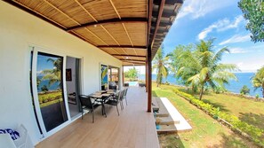 A covered outdoor area with a wooden deck, a dining table with chairs, and a view of the ocean and palm trees.