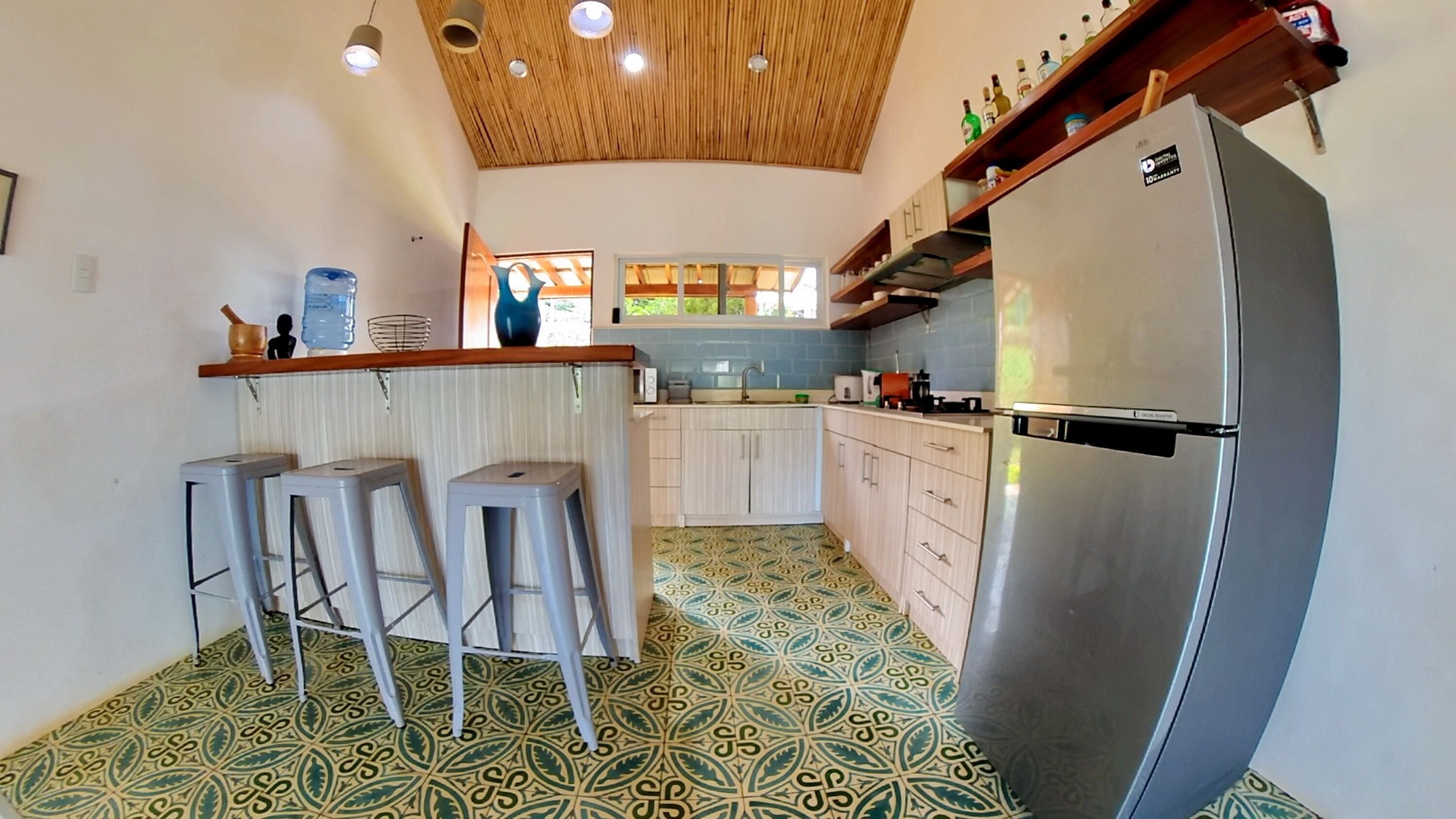 A compact kitchen with a stainless steel refrigerator, a bar counter with stools, and patterned floor tiles.