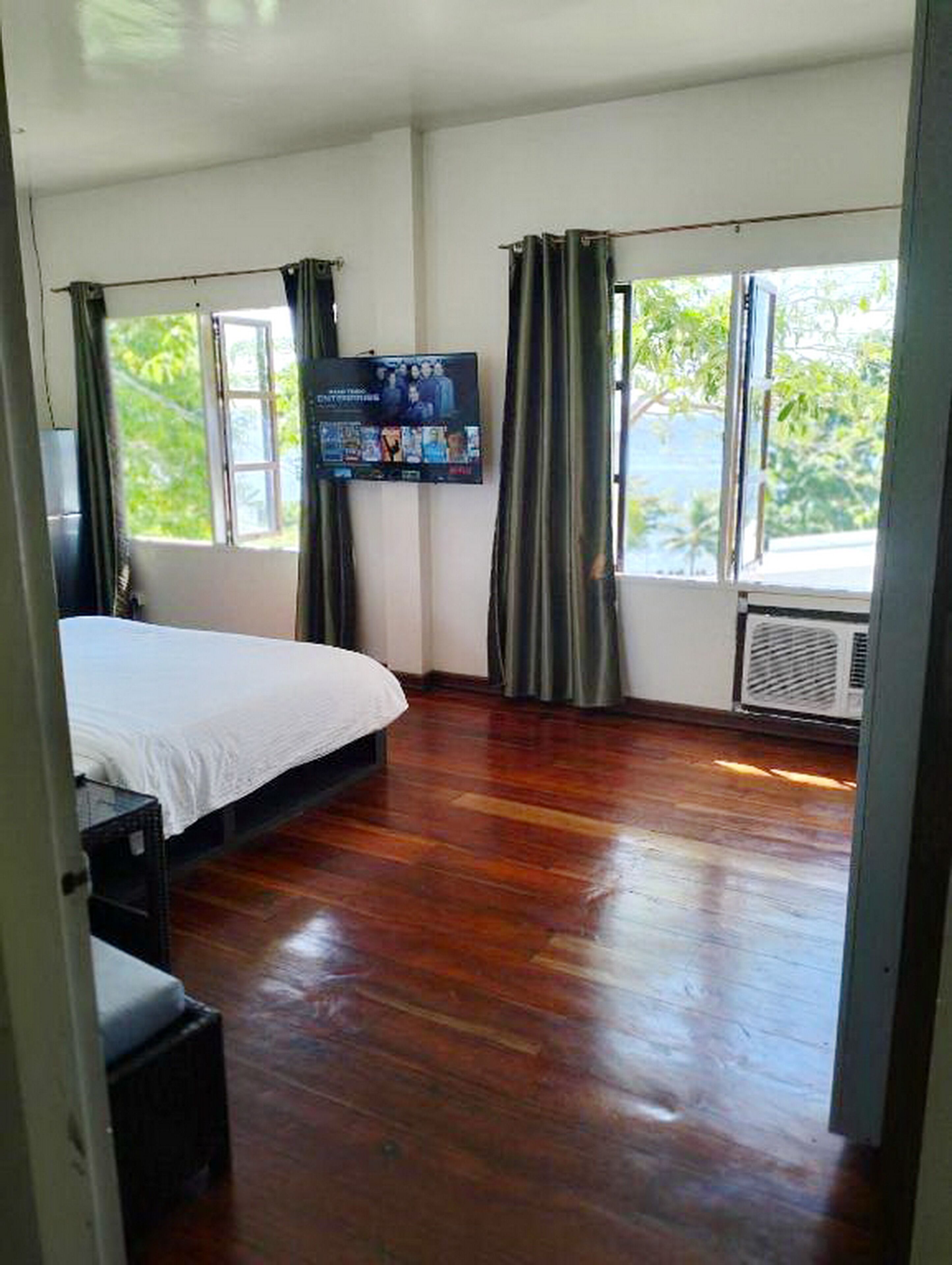 Quarto com cama, televisão fixada na parede e amplas janelas com cortinas.