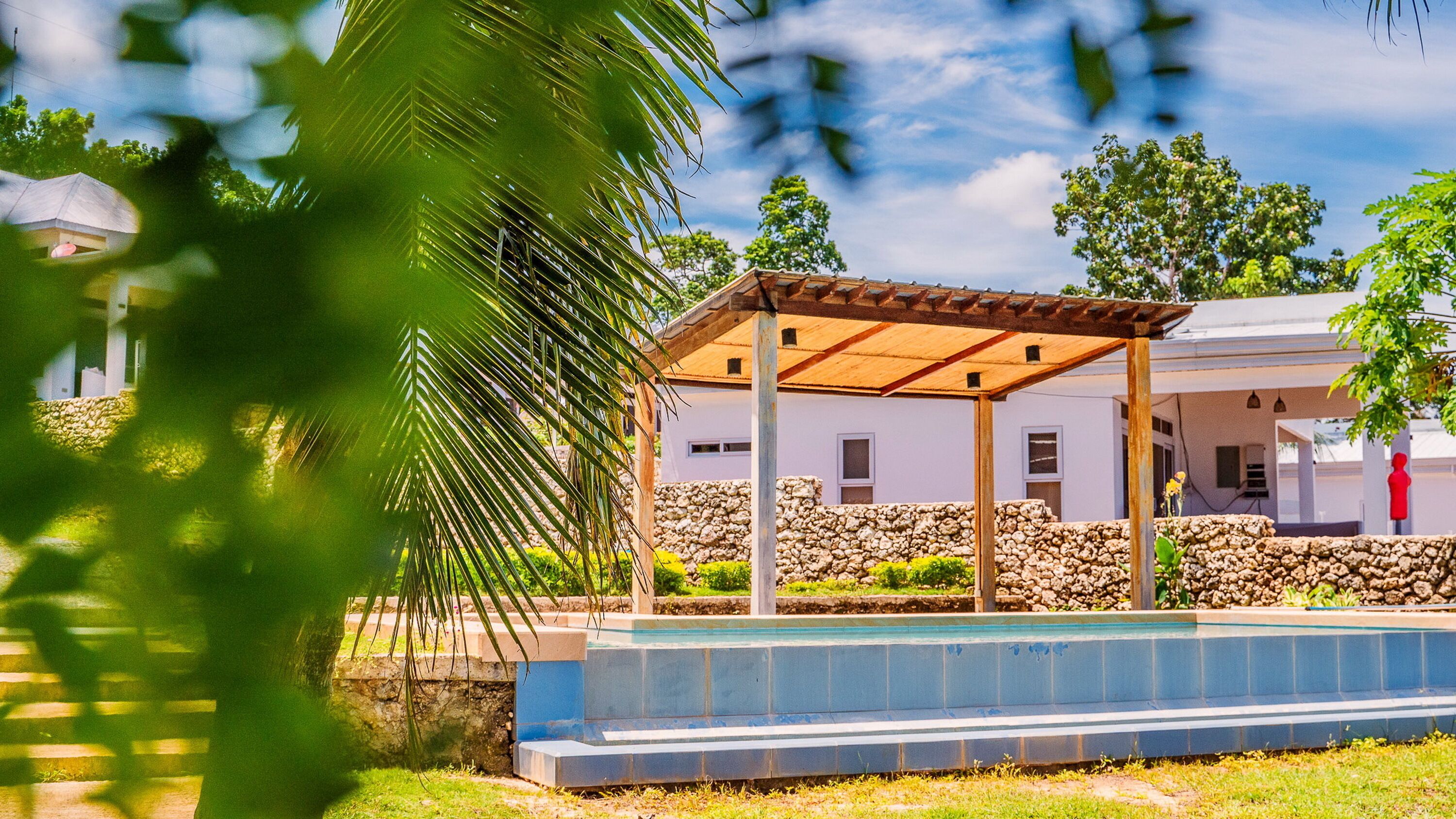 A covered outdoor seating area with a pool, stone walls, and a white building in the background.