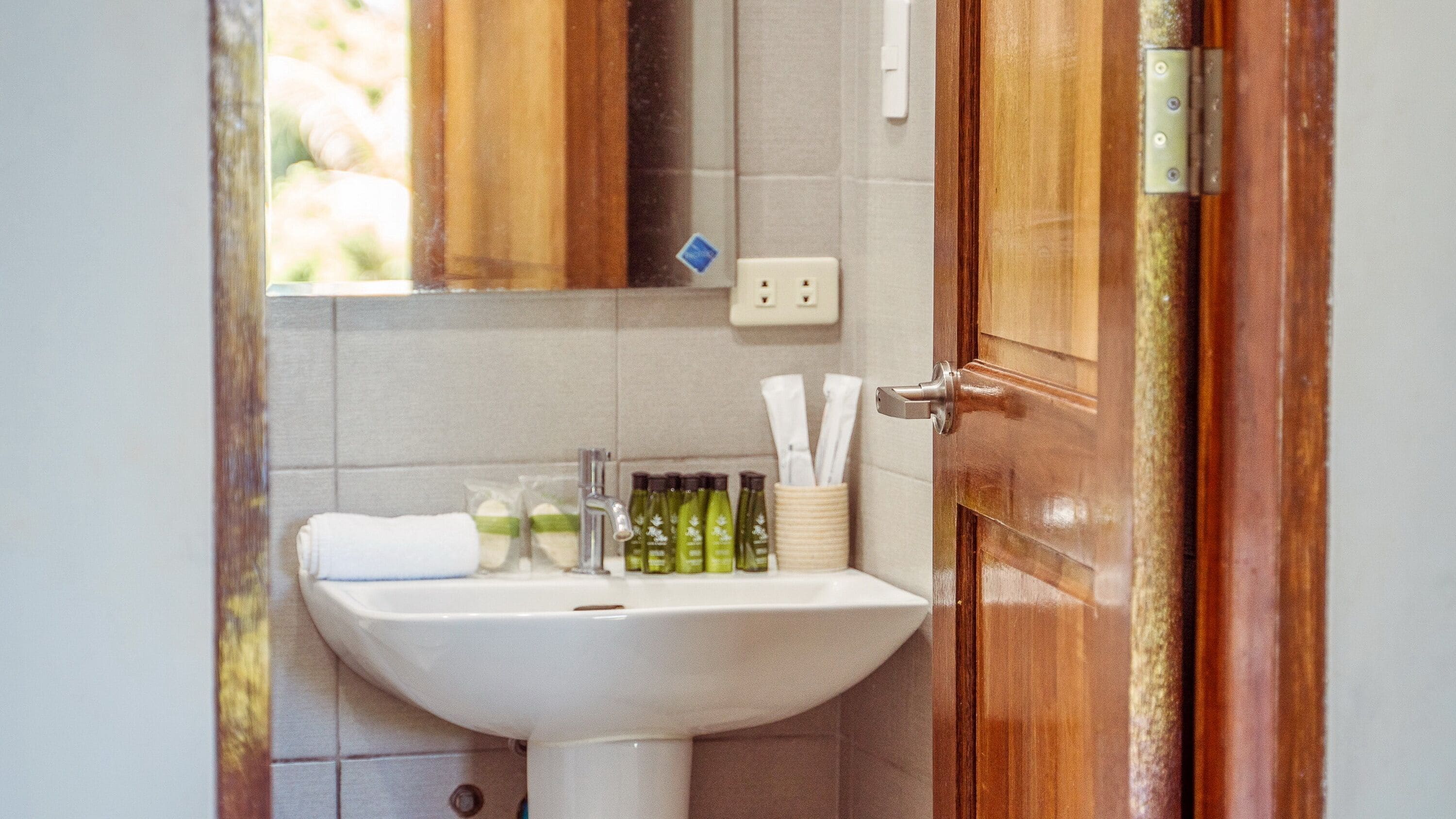A bathroom with a white sink, a mirror, and a wooden door. There are toiletries on the sink and a towel hanging on the wall.
