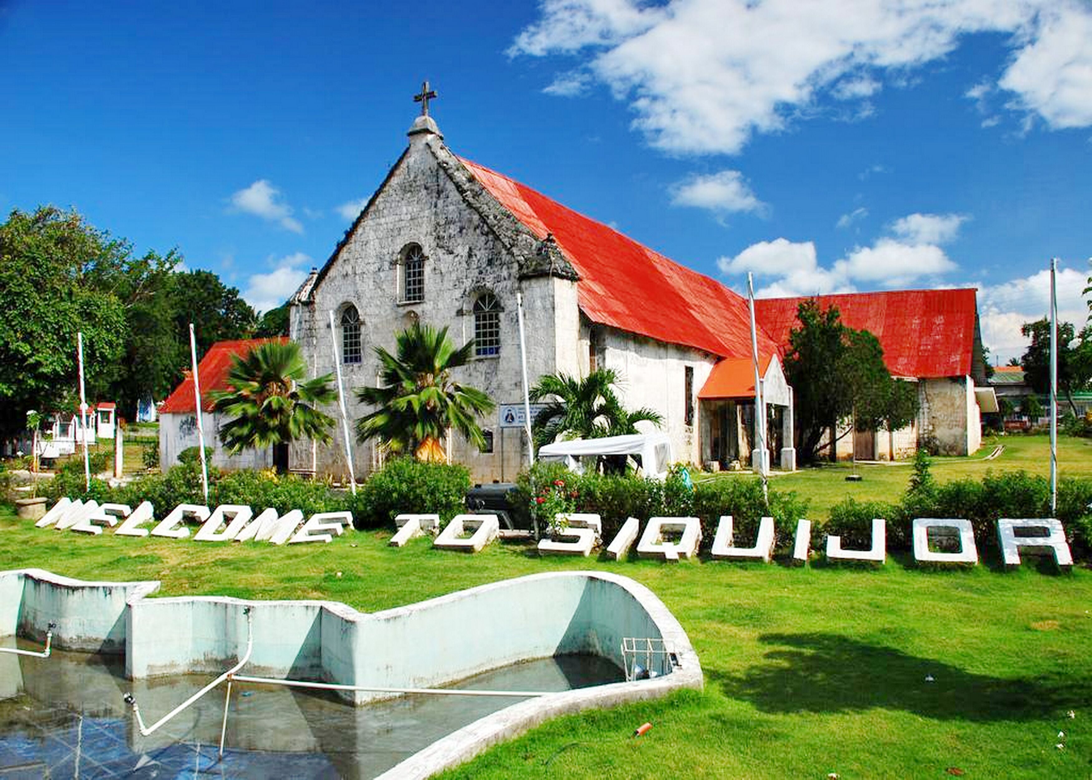 A church with a red roof and a cross on top, surrounded by greenery and a sign that reads "WELCOME TO SIQUIJOR".