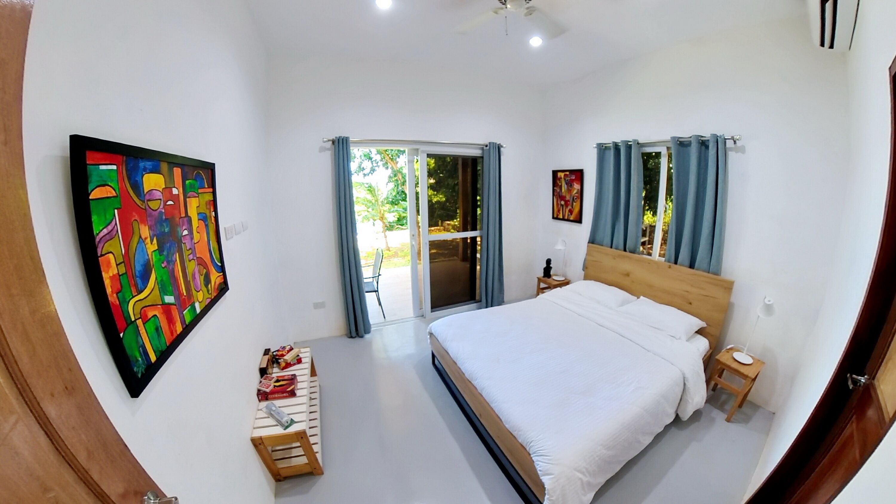 A bedroom with a bed, a nightstand, a chair, a table with books, a painting, and a sliding glass door to the outside.