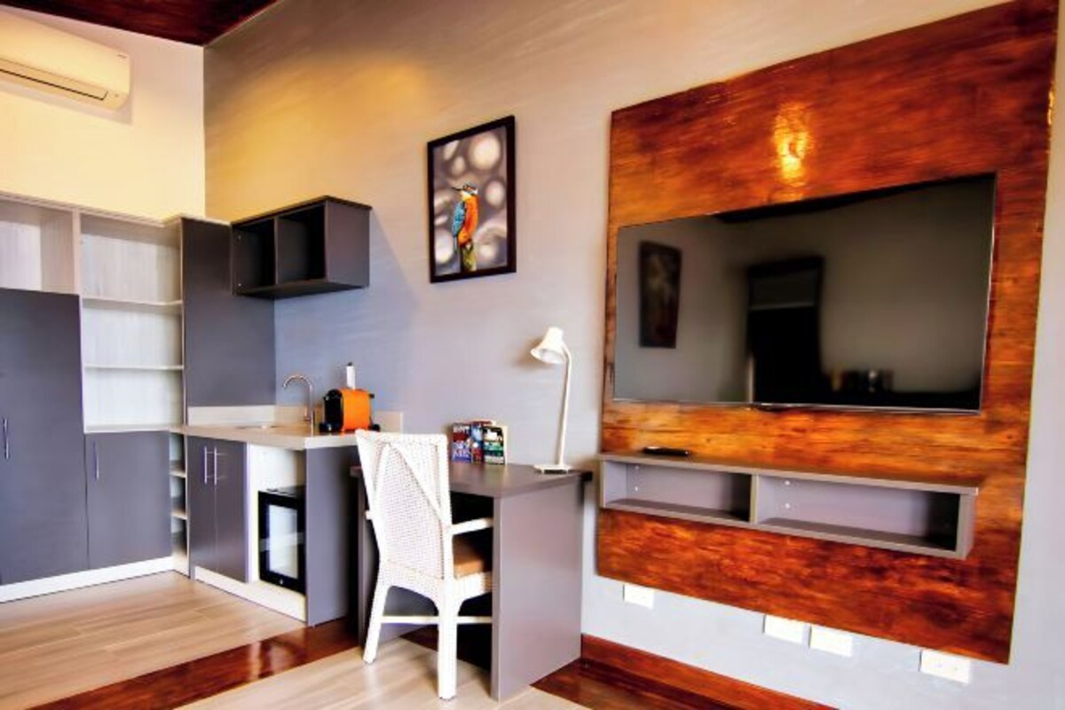 A compact kitchen with built-in cabinets, a white chair, a small sink, and a flat-screen TV mounted on a wooden accent wall.