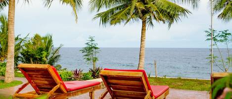 Two wooden lounge chairs with red cushions on a stone pathway, overlooking a body of water with palm trees in the background.