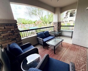 A balcony with wicker furniture, a glass table, and a view of a swimming pool and greenery.