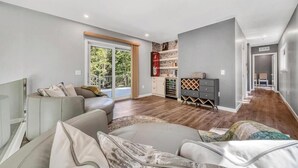 Een ruime woonkamer met een zithoek, een houten vloer, een schuifdeur naar een balkon en een inbouw wijnrek.