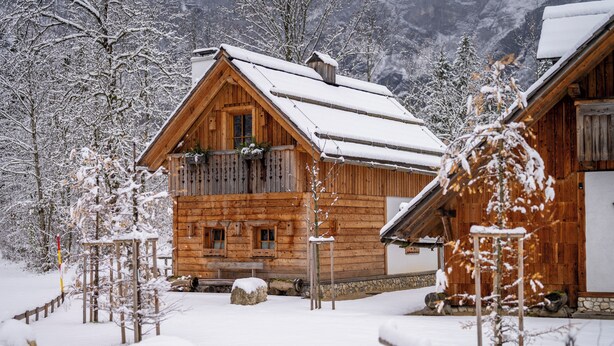 Una cabaña de madera con balcón, rodeada de árboles y montañas cubiertas de nieve.