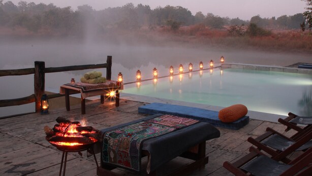 A serene outdoor pool area with a wooden deck, lounge chairs, a fire pit, and a row of lanterns along the pool's edge. - Saccharum Safari Lodge (Baihar)