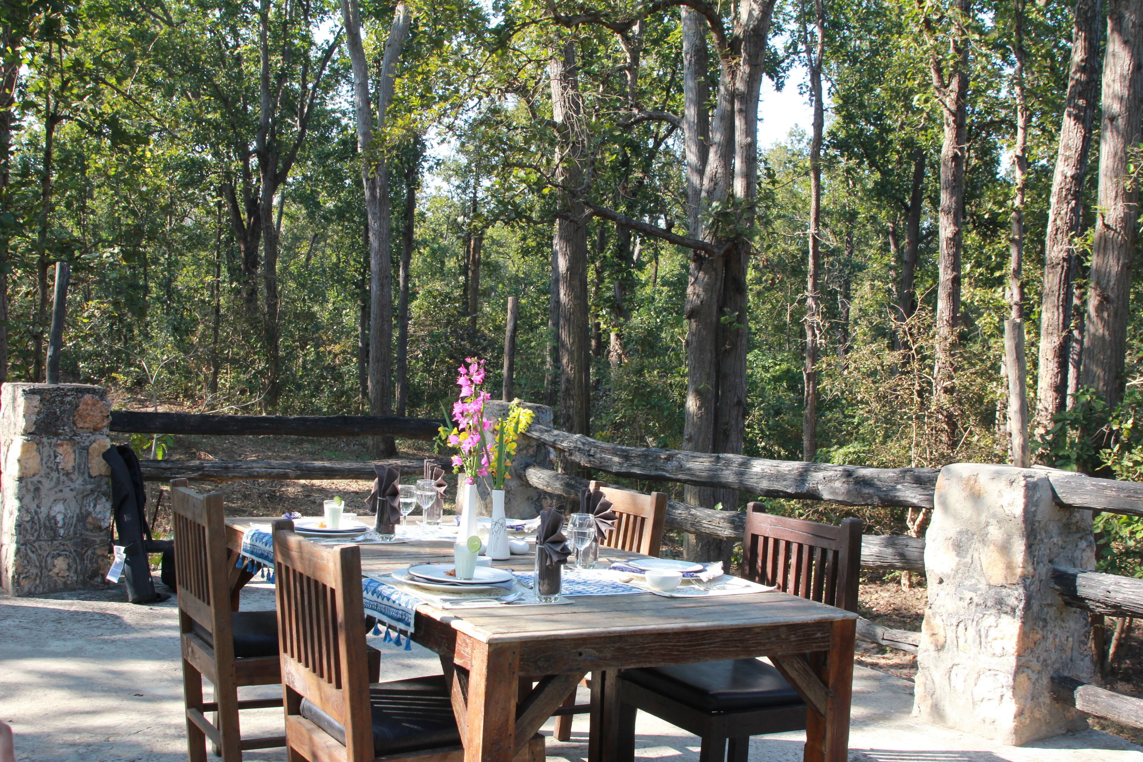 An outdoor dining area with a wooden table set for two, surrounded by chairs, in a forested setting.