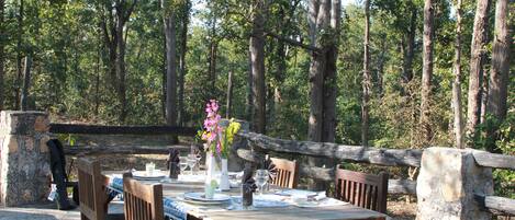 An outdoor dining area with a wooden table set for two, surrounded by chairs, in a forested setting.