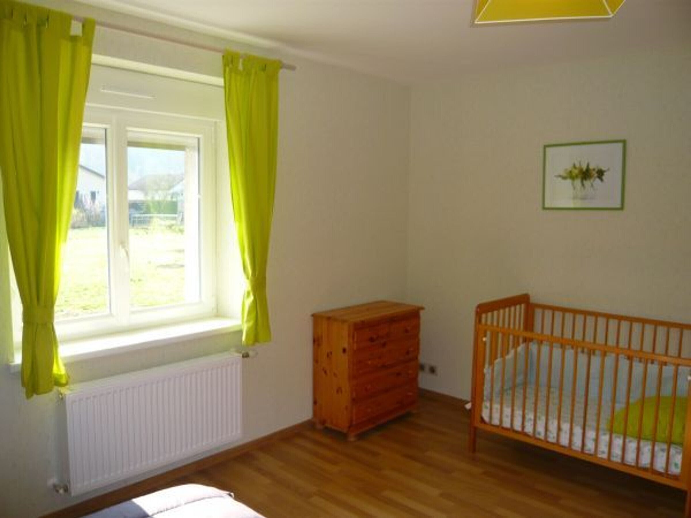 Een kamer met een houten vloer, een kinderbedje, een houten dressoir en een raam met gordijnen.