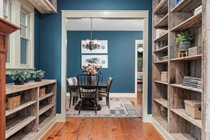 A dining area with a wooden table, chairs, and a chandelier. There are shelves on either side with books and plants. - Grit&Grace Ranch - Private Resort Style Pool & Spa (Austin)