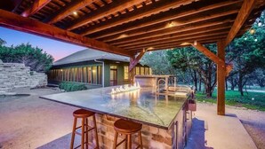 An outdoor kitchen area with a stone wall, a wooden pergola, and a bar with stools. - Grit&Grace Ranch - Private Resort Style Pool & Spa (Austin)