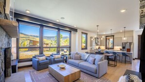 A spacious living room with a large sofa, a coffee table, and a fireplace. There are large windows offering a view of the outdoors. - Pet-Friendly Mountain Escape | Private Firepit | Near Trails + Adventure (Silverthorne)