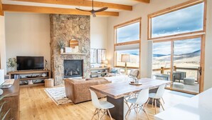 Un salón espacioso con chimenea de piedra, vigas de madera, un televisor grande y una zona de comedor con vistas al paisaje desértico.