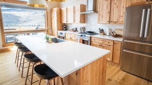 Una cocina moderna con armarios de madera, una isla grande con taburetes de bar, electrodomésticos de acero inoxidable y vistas a montañas nevadas a través de la ventana.