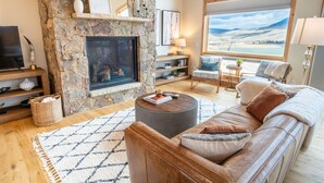 Una sala de estar acogedora con chimenea de piedra, un sofá de cuero y un amplio ventanal con vistas a un paisaje pintoresco.