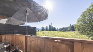 Une terrasse en bois, dotée de mobilier d’extérieur, et offrant une vue sur un champ verdoyant et des montagnes en arrière-plan.