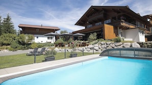 Vue sur un bâtiment de style chalet, avec une grande piscine, un espace salon extérieur et un jardin bien entretenu, le tout vu depuis le bord de la piscine.