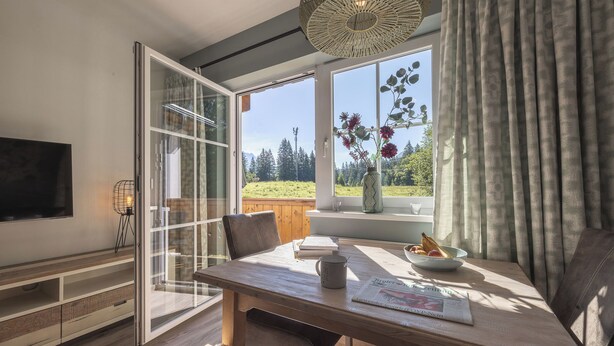 Un salon moderne avec une table à manger en bois, un téléviseur fixé au mur et une grande fenêtre donnant sur un paysage verdoyant.