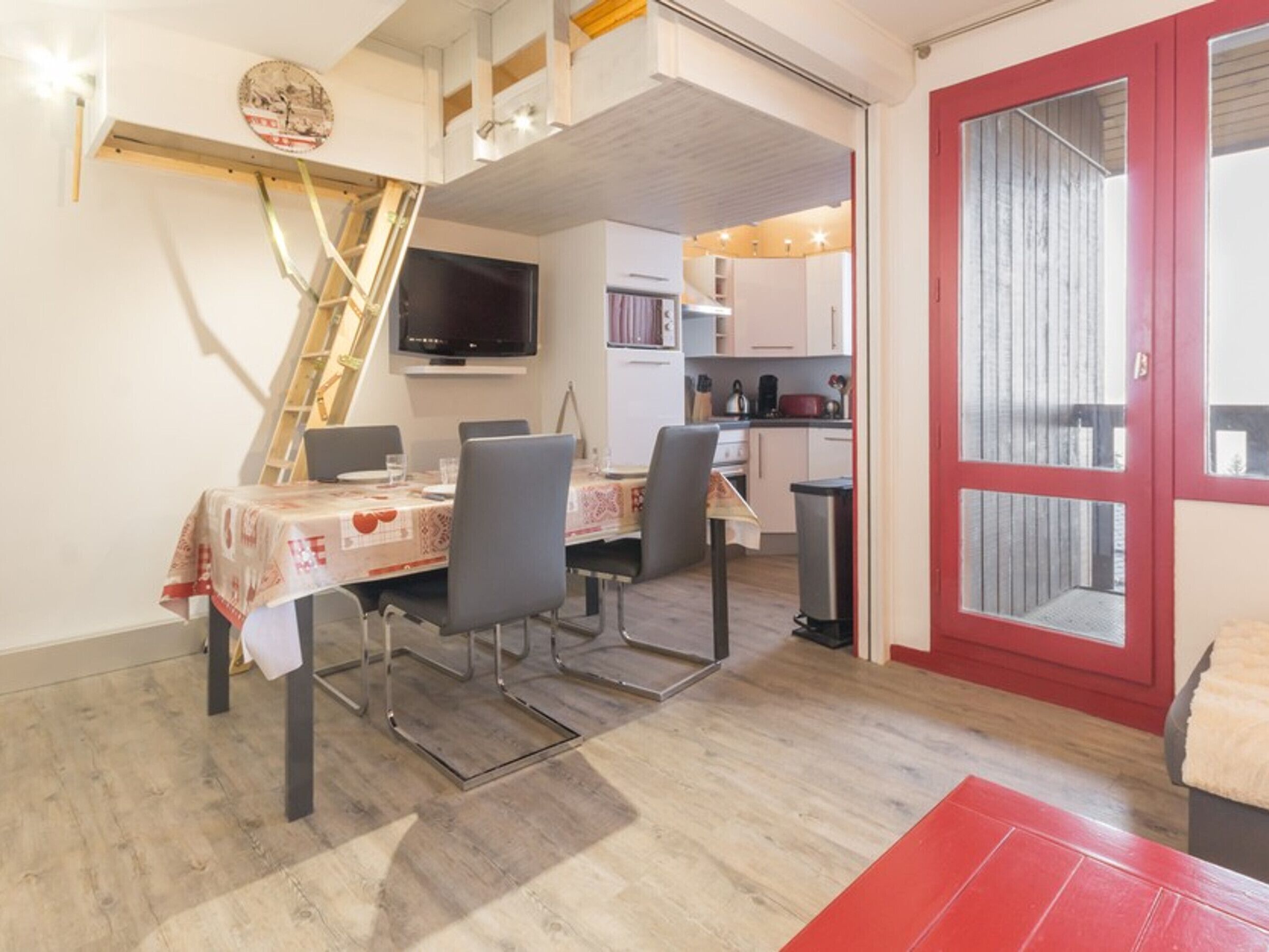 A dining area with a table set for four, a TV mounted above the table, and a red door leading to a balcony.