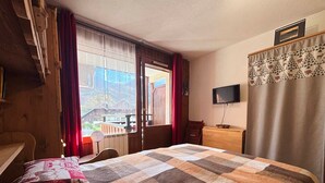 A hotel room with a large bed, a wooden dresser, a television, and a window with curtains. - Cozy studio in village center, pets allowed, private parking (Samoëns)
