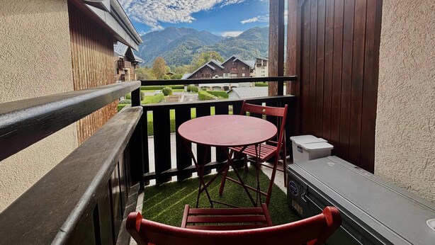 A balcony with a red table and chairs, a small grill, and a view of mountains and houses. - Cozy studio in village center, pets allowed, private parking (Samoëns)