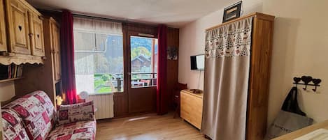 A cozy room with a wooden floor, a patterned sofa, a wooden cabinet, a television, and a window with curtains.