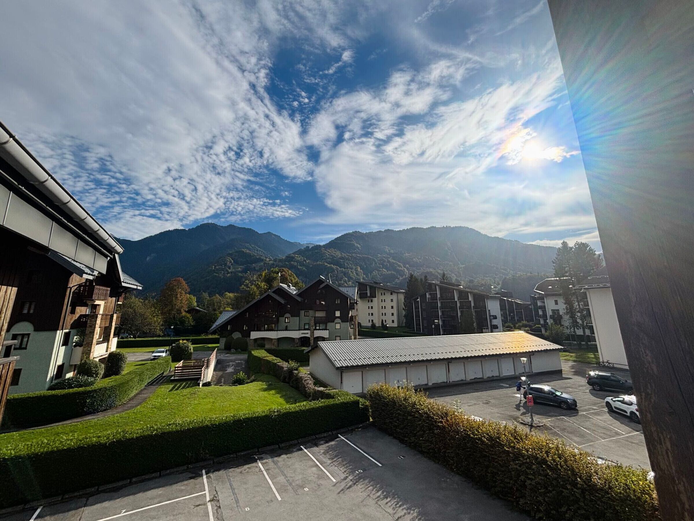 Blick auf einen Parkplatz mit mehreren Autos, ein Gebäude mit schrägem Dach und Berge im Hintergrund.