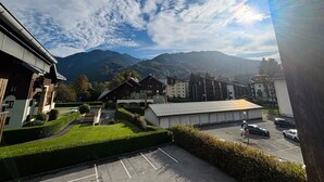 A view of a parking lot with several cars, a building with a sloped roof, and mountains in the background. - Cozy studio in village center, pets allowed, private parking (Samoëns)
