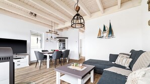 Ein modernes Wohnzimmer mit einer Couch, einem Couchtisch und einem Fernseher. Es gibt einen Essbereich mit Tisch und Stühlen. Der Raum hat Holzböden und eine hohe Decke mit sichtbaren Balken.